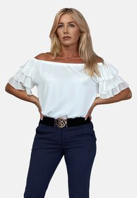 Biała bluzka na jedno ramię z falbankowymi rękawami, zestawiona z ciemnymi, wzorzystymi spodniami. Bluzka charakteryzuje się gładką teksturą i luźnym krojem.