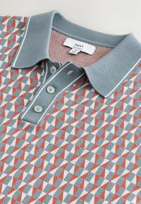 Collet et patte de boutonnage d'un polo bleu moyen avec un motif géométrique de triangles rouges, blancs et bleus et une étiquette de la marque "Next".