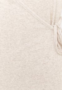 Tissu beige clair à côtes avec une texture douce, présentant un design croisé et un détail de lien sur le côté.