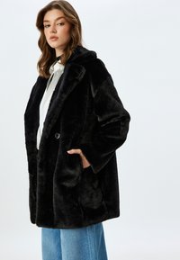 Sorte faux fur frakke med bred krave, enkelt knaplukning og sidelommer. Modellen har løstsiddende denimjeans og en hvid skjorte på.