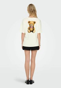 Crème kleurige oversized t-shirt met een grote teddybeerafbeelding met een roze strik op de rug, gecombineerd met zwarte shorts.