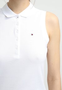 Vit ärmlös pikétröja med texturerad tyg, knappslå och en liten broderad logotyp i röd och blå på vänster bröst.