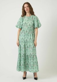 Robe maxi à motif floral vert avec manches courtes bouffantes, taille froncée et jupe à volants. Talons noirs complètent le look.