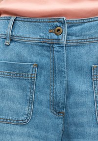 Plano detalle de unos vaqueros de mezclilla azul con botón metálico y bolsillos delanteros, llevados con una camiseta de tejido acanalado de color salmón.