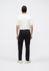 Jasnobeżowa koszula z krótkim rękawem, czarne spodnie i kremowe trampki. Włosy z charakterystycznym warkoczem, delikatny okrągły dekolt, widok od tyłu.