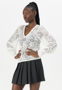 Witte, semi-doorzichtige pullover met een V-hals en lange mouwen, versierd met een ingewikkeld circulair patroon. Gecombineerd met een geplooide zwarte rok.