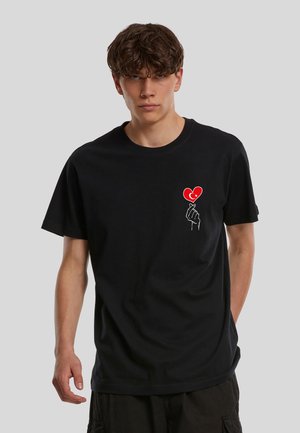 Jeune homme portant un t-shirt noir avec une main blanche tenant un cœur rouge orné d'un croissant de lune et d'une étoile sur la poitrine.
