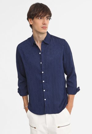 Marineblaues Hemd mit Knöpfen, strukturiertem Muster, langen, bis zu den Ellenbogen gerollten Ärmeln und weißen Knöpfen, kombiniert mit hellen Hosen.