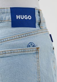 Jeans din denim de un albastru deschis cu o patch de piele albastră distinctă etichetată "HUGO." Prezintă cusături portocalii și o buzunar din spate cu logo embosat.