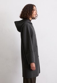 Grauester Wollmantel mit Kapuze, der über einen Knopfverschluss und zwei Seitentaschen verfügt. Das Design ist lang mit einer lässigen Silhouette und subtiler Textur.