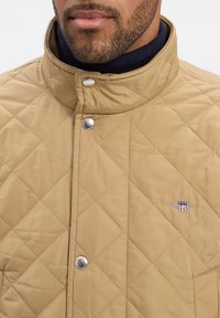 Beige vuoritettu takki, jossa on korkea kaulus, hopeiset napit ja brodeerattu logo rinnassa. Valmistettu sileästä ja kestävästä kankaasta.