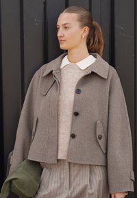 Pruun villaseguga lühike mantel kraega, mustade nuppude ja küljetaskutega, kantud beeži kampsuni ja triibuliste pükste peal. Roheline kott kaasa.