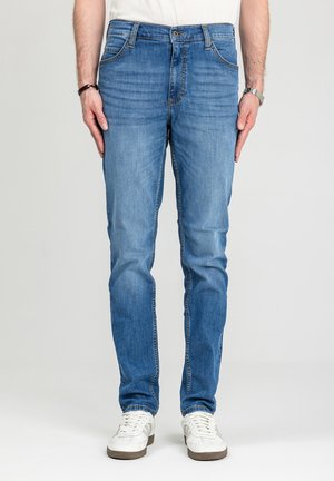 Mężczyzna noszący niebieskie dżinsy o dopasowanym kroju i białe trampki, stojący z rękami swobodnie opuszczonymi wzdłuż ciała na jednolitym tle.