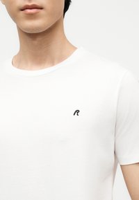 Ung mann som har på seg en enkel hvit T-skjorte med korte ermer og en liten svart bokstav "R" brodert på venstre bryst.