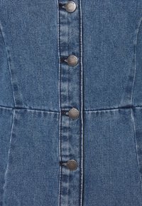 Blåt denimstof med lodret søm og fire metalknapper med indgraveret "JOY DENIM" på et tøjs forreste midte.