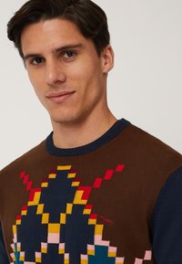 Gebreide trui met een geometrisch patroon in bruin, rood, geel en blauw, met een donkerblauwe halslijn. Subtiel logo in het ontwerp.