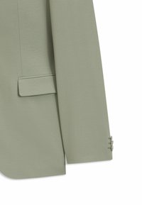 Rukáv saka v barvě šalvějové zeleně s třemi knoflíky u manžety a kapsou s klopou na těle proti bílému pozadí.