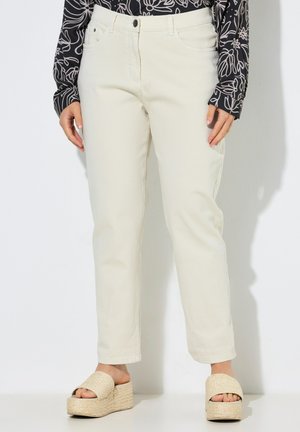 Vrouw met crème rechte broeken, zwart-wit patroon lange mouwen top, en beige plateau sandalen.