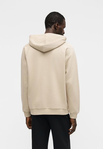 Homme portant un sweat à capuche beige et un pantalon noir, vu de dos sur un fond uni clair.
