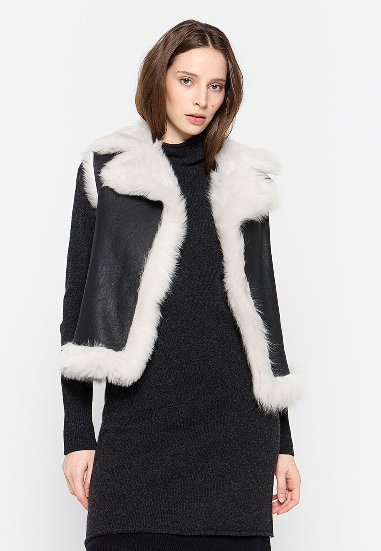 Schwarze, ärmellose Lederweste mit weißem Fellbesatz. Sie verfügt über ein minimalistisches Design und eine glatte Textur, die über einem dunklen Strickkleid getragen wird.