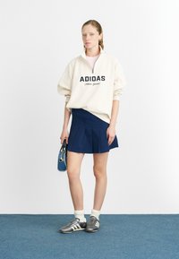 Felpa crema con logo "ADIDAS", gonna plissettata blu, scarpe da ginnastica grigie e calzini bianchi; regge una borsa blu con tracolla.