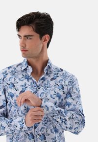 Camicia blu a fantasia floreale con colletto aperto, polsini con bottone e una texture di tessuto leggera. Presenta diverse sfumature di blu e bianco.
