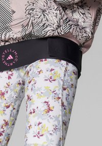 Cintura fitness nera con logo rosa, abbinata a leggings a fantasia floreale con colori giallo, viola e grigio. Texture del tessuto liscia.