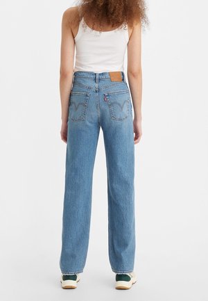 Vrouw met krullend haar die een wit tanktop draagt, hoge rechte blauwe jeans en witte sneakers, staand met de rug naar voren.