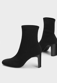 Svarta sockboots i medellång längd med rundade tår och blockklackar, tillverkade av slät tyg, visade från sidan och bakifrån på en vit bakgrund.