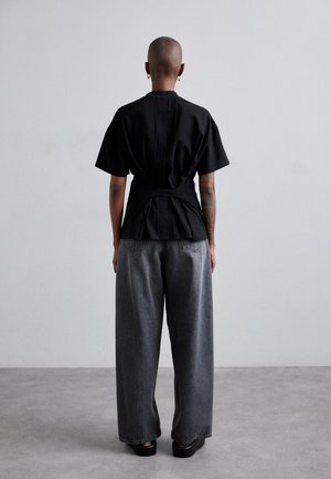 Personne avec la tête rasée et le bras droit tatoué, portant un haut noir à manches courtes et un jean gris à jambes larges, debout devant un fond clair uni.