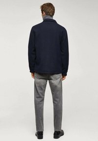 Navy wool blend jacket with a high collar, worn over gray jeans. The jacket features a textured finish and side pockets. Black shoes are visible.