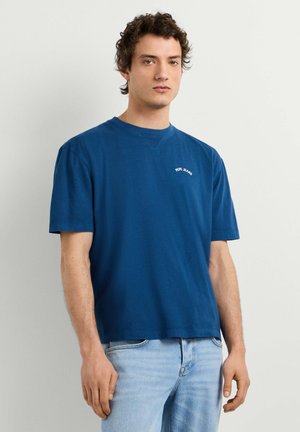 Ung mand med krøllet hår iført en blå Pepe Jeans t-shirt og lyseblå jeans, stående foran en ensartet lys baggrund.