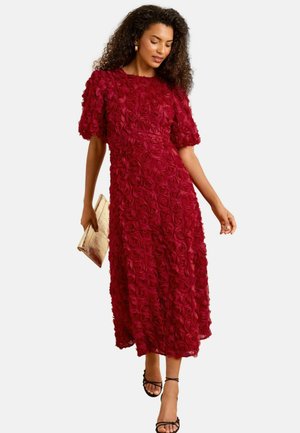 Red rose-patterned midi dress with puff sleeves. Texture features layered floral embellishments. Model holding a gold clutch and wearing black sandals.