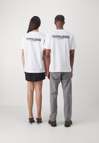 Camisetas de algodón blancas con el logo de 'Tommy Jeans' impreso en rojo y negro en la parte posterior. Combinadas con jeans grises y una falda negra.