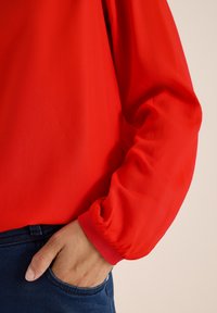 Rote Bluse aus weichem, transparentem Stoff mit elastischen Bündchen. Sie hat eine lockere Passform und wird mit dunkler Denim-Jeans kombiniert.