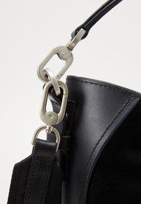 Detalj av en svart läderhandväska med en metallisk, silverfärgad spänne, slät yta och sydda kanter. Inkluderar en tygrem som accent.