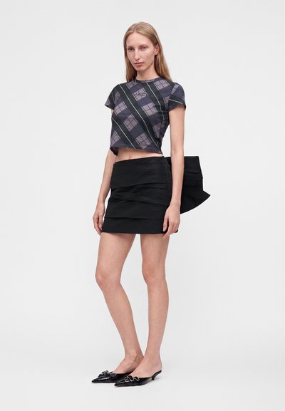 Jeune femme portant un crop top à carreaux noirs et violets, une mini-jupe noire à volants sur le côté et des chaussures slingback noires pointues.
