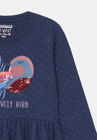 Navyblaues Langarmkleid mit einem aus Pailletten bestehenden Vogel-Graphic, mit gepunkteter Textur und gerafftem Bund. Text lautet "Lovely Bird."