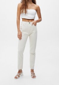 Top cortado blanco sin tirantes combinado con jeans rectos de tiro alto en color blanco roto. Sandalias blancas de tacón alto con tiras completan el atuendo.