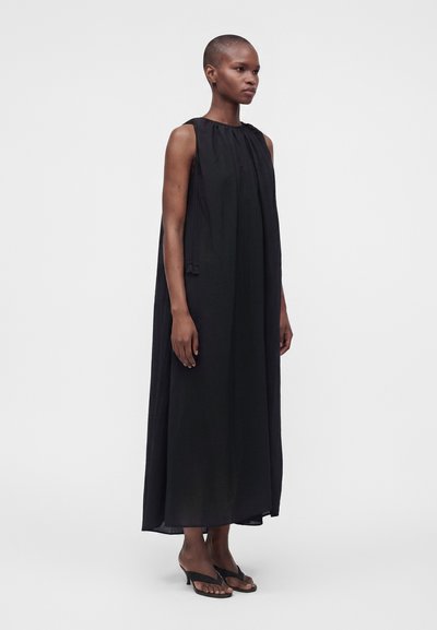 Femme aux cheveux courts portant une robe maxi noire ample sans manches et des sandales à talons noires, debout devant un fond clair uni.