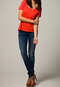 Kvinna står med korslagda ben, iklädd en röd t-shirt, blå jeans och bruna högklackade skor, och hållande sin vänstra arm med höger hand.