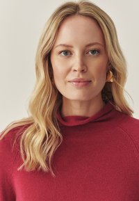 Czerwony sweter z dzianiny o wysokim dekolcie, z ryflowaną fakturą. Modelka ma falowane blond włosy i nosi złote kolczyki. Neutralne tło.