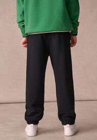 Fekete polár jogging nadrág elasztikus mandzsettával, fehér sportcipővel párosítva. Zöld pulóver látható fent, kontrasztos belső réteggel.