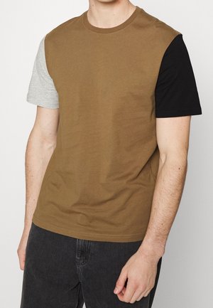 Mann trägt ein kurzärmliges braunes T-Shirt mit einem grauen Ärmel und einem schwarzen Ärmel, kombiniert mit dunkler Jeans, vor weißem Hintergrund.