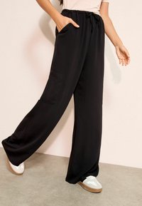 Fekete, széles szárú nadrág elasztikus derékrésszel és húzózsinórral, oldalzsebekkel és sima, könnyű anyagból. Sportcipővel kiegészítve.