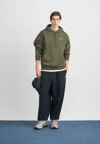 Ελιάς πράσινη φούτερ με κουκούλα και μπροστινή τσέπη, συνδυασμένη με φαρδιά μαύρα παντελόνια και γκρίζα αθλητικά παπούτσια. Κρατώντας ένα ναυτικό καπέλο. Απλός σχεδιασμός.