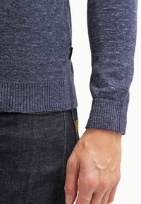 Pull en laine bleu à finition texturée, avec des poignets et un ourlet côtelés ; présenté à côté d'un jean en denim foncé et d'une main reposant sur le bord.