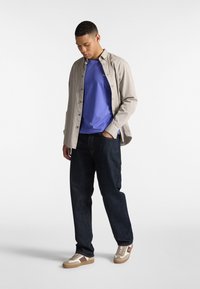 Outfit casual composto da una camicia button-up beige chiaro, una t-shirt viola, jeans in denim scuro e sneakers beige con accenti marroni.