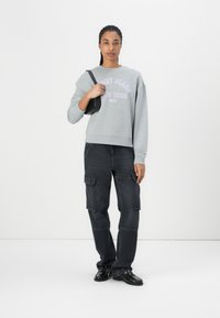 Felpa grigia con testo viola e collo rotondo, abbinata a pantaloni cargo scuri con tasche multiple. Borsa nera e scarpe.