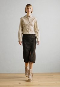 Chemise beige courte avec taille froncée, jupe noire avec fente à l'avant, et bottines à paillettes beiges. Sol en bois et arrière-plan uni.